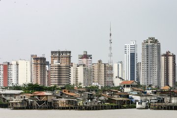 Urban landscape featuring a mix of modern high-rise buildings and informal riverside shacks, with a prominent communication tower in the background.