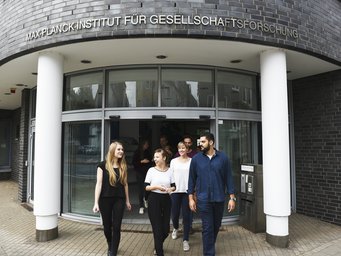 Das Foto stellt den Eingangsbereich des MPIfG vor. Mehrere Individuen der Gattung Homo sapiens intellectualis treten heraus, andere befinden sich noch unmittelbar an der Schließtür. Sie zeigen sich einander freundlich zugewandt. Offenbar haben sie den Entschluss gefasst, gemeinsam einen Gang hinaus zu unternehmen.