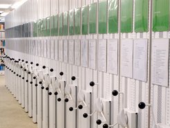 A series of mobile library shelves, each fitted with crank handles for manual adjustment. Above them, documents and green folders are displayed, indicating catalog information.