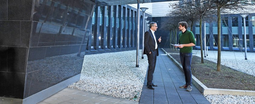 Zwei Männer unterhalten sich im Freien eines modernen Gebäudekomplexes mit Gehwegen und Bäumen.