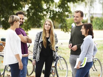 A mixed group of five people stands in a park, engaged in conversation. Two bicycles are visible in the background, hinting at a casual meeting or a break during a bike ride. The setting is relaxed, with trees providing shade.