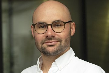 The photo shows an individual of the species Homo sapiens intellectualis named Björn Bremer. A middle-aged man with a clean-shaven head and faint signs of attempted beard growth. The individual wears round, orthopedically fitted glass windows on his nose, which generally serve the purpose of ensuring better visibility in the thicket of reality.